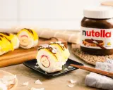 Süße Sushi-Rollen mit Milchreis als Basis, gefüllt mit Banane und Erdbeere, garniert mit gelbem Fruchtpüree, Schokodrizzle und Kokoschips. Daneben eine Nutella-Packung.