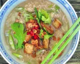Khao tom, ein herzhafter Reissuppe mit gebratenem Tofu, Frühlingszwiebeln und Kräutern, serviert in einer blauen Schüssel mit Essstäbchen.