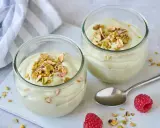 Zwei Gläser mit cremiger Pistazienmousse, garniert mit gehackten Pistazien. Ein Löffel und Himbeeren liegen daneben.