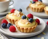 Zwei köstliche Vanille-Tartelettes mit frischen Beeren und Mandelsplittern, serviert auf einem Teller mit einer Kaffeetasse im Hintergrund.