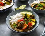 Schüssel mit Reis und rotem Thai Curry mit Rindfleisch, Zucchini und Paprika, garniert mit Limettenspalten und Koriander.