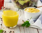 Gelbe Vinaigrette in einem Glasbehälter, bereit für einen frischen Salat. Daneben eine Schüssel Senf und ein Salat im Hintergrund.