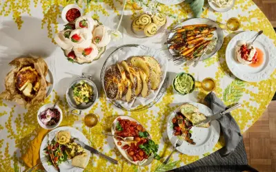 Tisch voller Oster-Essen-Ideen: Meringues mit Erdbeeren, Brot, Karotten, Quiche und mehr auf gelb-weißem Tischtuch.