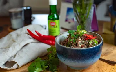 Schüssel mit würzigem Larb Moo, garniert mit frischen Kräutern und Chilis. Daneben liegen rote Chilis und eine Flasche Sauce.