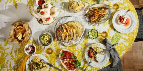 Tisch voller Oster-Essen-Ideen: Meringues mit Erdbeeren, Brot, Karotten, Quiche und mehr auf gelb-weißem Tischtuch.