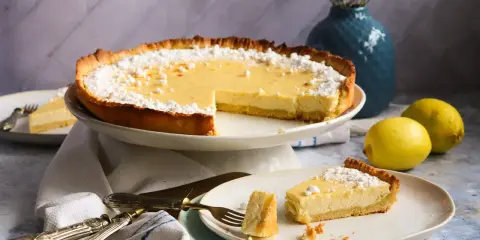 Eine köstliche Zitronen-Tarte, bestäubt mit Puderzucker, steht auf einem Kuchenständer. Ein Stück liegt auf einem Teller, daneben zwei frische Zitronen.