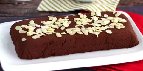 Dunkle, rechteckige marquise au chocolat, bestreut mit Mandelblättchen, auf einem weißen Teller.