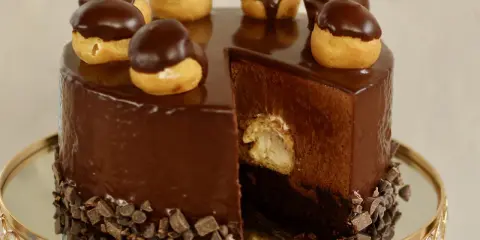 Eine glänzende Schokoladentorte, dekoriert mit kleinen Schokoladen-Profiteroles. Ein Stück ist herausgeschnitten und zeigt die cremige Füllung.