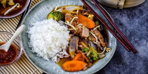 Reis mit einem herzhaften Stir-Fry-Gericht, das mit viel Gemüse und Fleisch in einer köstlichen Sauce serviert wird. Dazu gibt es eine kleine Schale mit scharfer Sauce.