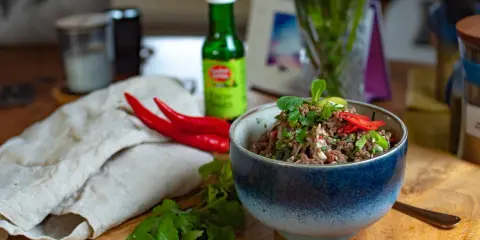 Schüssel mit würzigem Larb Moo, garniert mit frischen Kräutern und Chilis. Daneben liegen rote Chilis und eine Flasche Sauce.