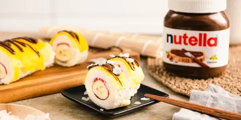 Süße Sushi-Rollen mit Milchreis als Basis, gefüllt mit Banane und Erdbeere, garniert mit gelbem Fruchtpüree, Schokodrizzle und Kokoschips. Daneben eine Nutella-Packung.