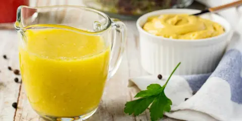Gelbe Vinaigrette in einem Glasbehälter, bereit für einen frischen Salat. Daneben eine Schüssel Senf und ein Salat im Hintergrund.