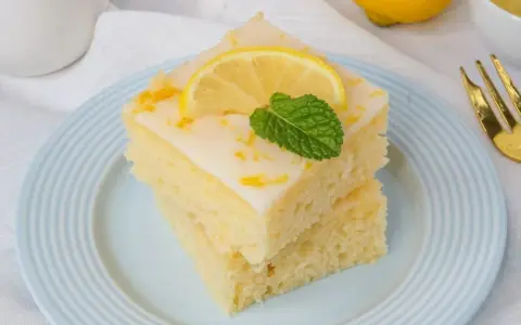 Zwei saftige Stücke weltbester Zitronenkuchen mit Joghurt, garniert mit Zitrone und Minze, auf einem blauen Teller.