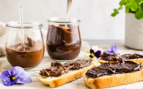 Zwei Gläser mit schoko creme, daneben Brotscheiben, üppig bestrichen mit der cremigen Schoko-Leckerei, verziert mit Veilchen.