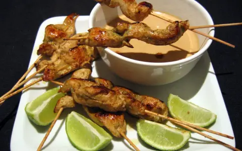 Saftige sate spiesse mit Erdnusssauce und Limettenspalten auf einem weißen Teller.