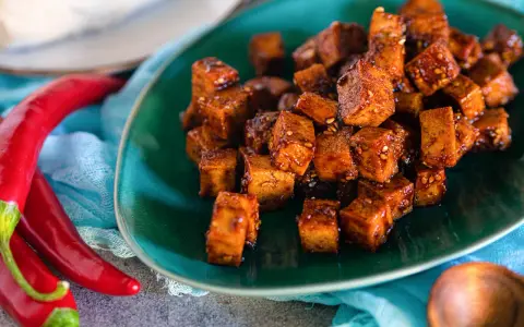Würzig gebratene Tofuwürfel auf einem grünen Teller, daneben rote Chilis. So schmeckt Tofu!