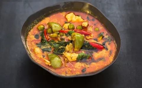 Ein dampfender rote thai curry mit Hühnchen, grünen Auberginen und roten Chilis in einer dunklen Schüssel.