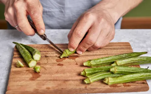 Hände schneiden Okra für ein leckeres Okra-Gericht. Frische grüne Schoten liegen auf einem Holzbrett.