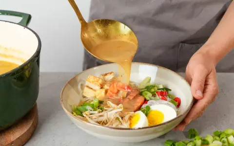 Eine goldene Schöpfkelle gießt eine cremige Brühe über Garnelen, Tofu, Ei und Sprossen in einer Schüssel. Ein köstliches Laksa Rezept wird zubereitet.