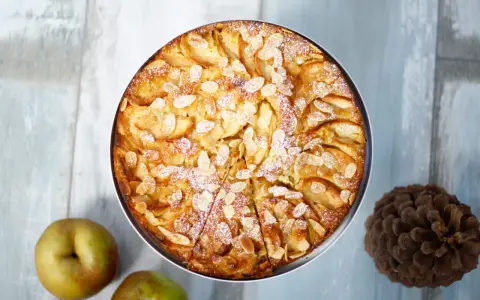 Ein goldbrauner bretonischer Apfelkuchen, bestreut mit Mandelblättchen und Puderzucker, neben Äpfeln und einer Kiefernzapfen.