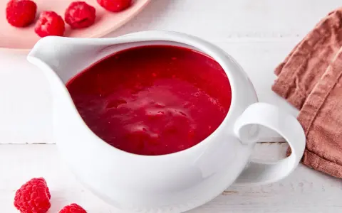 Himbeer coulis in einer weißen Soßenkelle, daneben frische Himbeeren und eine rosa Platte mit weiteren Früchten.