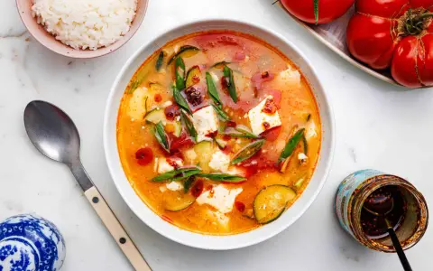 Eine dampfende Schüssel seidentofu suppe mit Zucchini und Frühlingszwiebeln, garniert mit Chiliöl. Daneben eine Schüssel Reis und ein Glas Chiliöl.