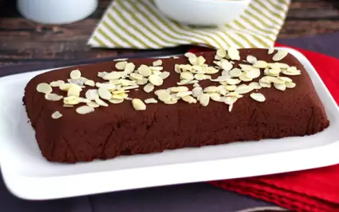 Dunkle, rechteckige marquise au chocolat, bestreut mit Mandelblättchen, auf einem weißen Teller.