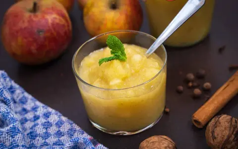 Ein Glas mit hausgemachtem compote de pommes, garniert mit Minze. Äpfel, Zimt und Nüsse im Hintergrund.
