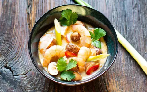 Eine Schüssel mit cremigem Tom Kha Gung, garniert mit Garnelen, Pilzen und Koriander, auf rustikalem Holz.