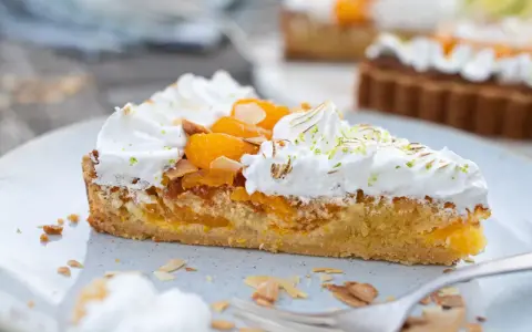 Ein Stück Mandarinen Tarte mit Baiserhaube, Mandelsplittern und frischer Zitronenschale bestreut.