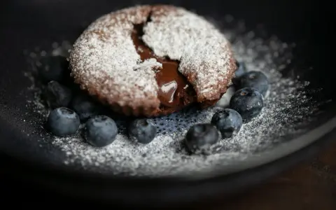 Ein saftiger Moelleux au Chocolat, bestäubt mit Puderzucker, umgeben von frischen Blaubeeren auf einem dunklen Teller.