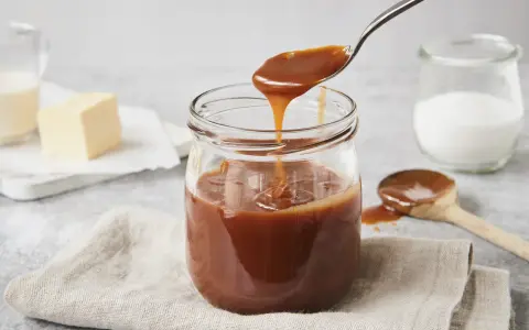 Ein Glas mit glänzender karamellsauce mit milch statt sahne. Ein Löffel hebt die cremige Sauce an.