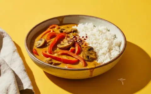 Schüssel mit Reis und rotem Curry, das mit Paprika und Pilzen verfeinert ist. Ein Hauch von Chiliflocken deutet an, dass dieses rote Curry scharf ist.