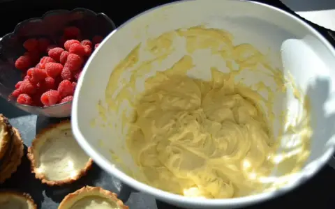 Frische Himbeeren warten neben leeren Tartelettförmchen und einer Schüssel mit cremiger Füllung auf die Fertigstellung der Himbeer Tartelettes.