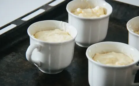 Vier weiße Förmchen mit luftiger Masse für ein Käsesoufflé stehen auf einem Backblech.