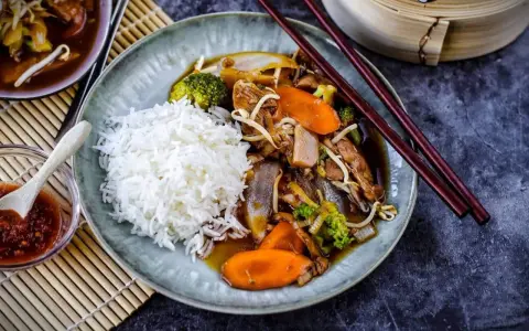 Reis mit einem herzhaften Stir-Fry-Gericht, das mit viel Gemüse und Fleisch in einer köstlichen Sauce serviert wird. Dazu gibt es eine kleine Schale mit scharfer Sauce.