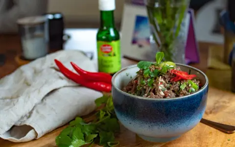 Schüssel mit würzigem Larb Moo, garniert mit frischen Kräutern und Chilis. Daneben liegen rote Chilis und eine Flasche Sauce.