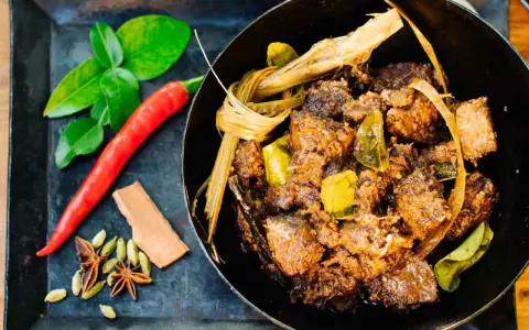 Ein dampfender Topf mit würzigem Beef Rendang, daneben frische Kaffirlimettenblätter, Chili und Gewürze.