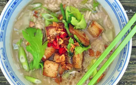 Khao tom, ein herzhafter Reissuppe mit gebratenem Tofu, Frühlingszwiebeln und Kräutern, serviert in einer blauen Schüssel mit Essstäbchen.