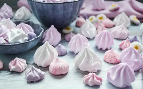 Zuckergebäck, was ist Meringue? Zarte, luftige Baisers in Rosa-, Lila- und Weißtönen, einige in einer Schüssel, andere verstreut.