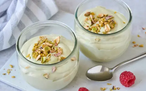 Zwei Gläser mit cremiger Pistazienmousse, garniert mit gehackten Pistazien. Ein Löffel und Himbeeren liegen daneben.