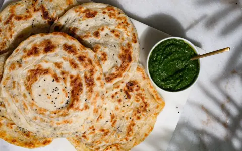 Fluffiger Roti Canai mit schwarzen Sesamkörnchen, daneben eine Schale mit grüner Chutney-Sauce.