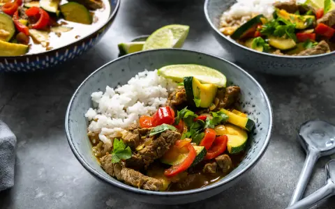 Schüssel mit Reis und rotem Thai Curry mit Rindfleisch, Zucchini und Paprika, garniert mit Limettenspalten und Koriander.