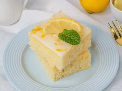 Zwei saftige Stücke weltbester Zitronenkuchen mit Joghurt, garniert mit Zitrone und Minze, auf einem blauen Teller.