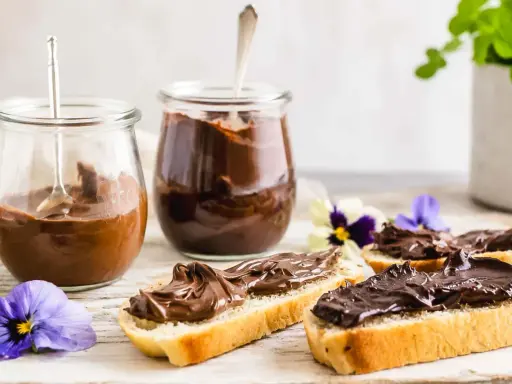 Zwei Gläser mit schoko creme, daneben Brotscheiben, üppig bestrichen mit der cremigen Schoko-Leckerei, verziert mit Veilchen.