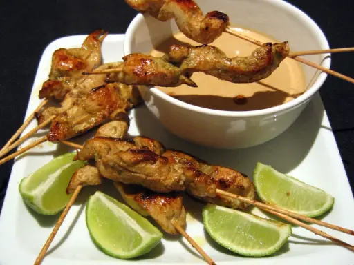 Saftige sate spiesse mit Erdnusssauce und Limettenspalten auf einem weißen Teller.