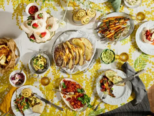 Tisch voller Oster-Essen-Ideen: Meringues mit Erdbeeren, Brot, Karotten, Quiche und mehr auf gelb-weißem Tischtuch.
