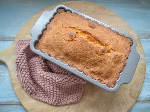 Frisch gebackener, goldbrauner quatre quarts in einer Kastenform, daneben ein rosafarbenes Stricktuch.