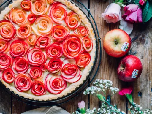 Ein wunderschöner Apfelrosen Kuchen, verziert mit zarten Apfelscheiben, die wie Rosen aussehen. Daneben liegen frische Äpfel und Blumen.