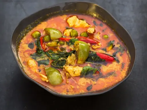 Ein dampfender rote thai curry mit Hühnchen, grünen Auberginen und roten Chilis in einer dunklen Schüssel.